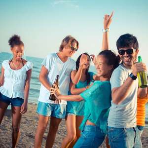 Smilende ungdom drikker og danser rygg mot rygg på en strand med et surfebrett i bakgrunnen (Foto)