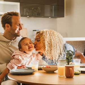 En mor kysser sitt barn på kinnet mens barnet sitter på farens fang ved frokostbordet (Foto)