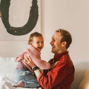 Far sitter i sofa og leker med en baby mens begge ler og smiler (Foto)