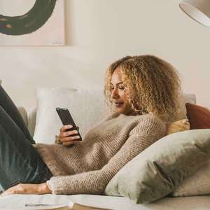Kvinne ligger i sofaen med mobiltelefonen, mens en åpen bok ligger på bordet ved siden av (Foto)