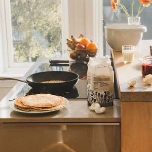 En tallerken med nystekte pannekaker står på en kjøkkenbenk, sammen med et par knuste egg, en pakke hvetemel og et åpent norgesglass med syltetøy - i bakgrunnen står stekepannen på platen men en pannekake på vei og en kurv med frukt ved siden av (Foto)