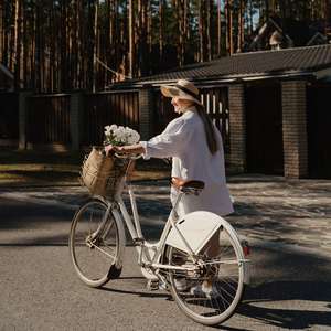 Kvinne i hvitt triller en hvit sykkel med en kurv blomster foran gjennom skogen (Foto)