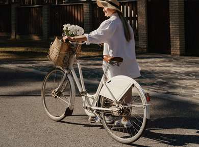 Kvinne i hvitt triller en hvit sykkel med en kurv blomster foran gjennom skogen (Foto)