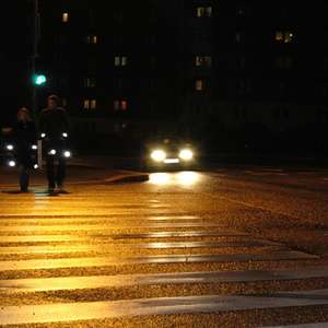 To personer med refleks krysser veien i mørket mens en bil kommer kjørende i bakgrunnen (Foto)