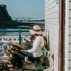 To mennesker sitter på en benk ved sjøen inntil en husvegg (Foto)