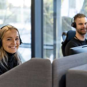 To ansatte på Frende sitt kundesenter snur seg smilende mot kamera fra pultene sine (Foto)