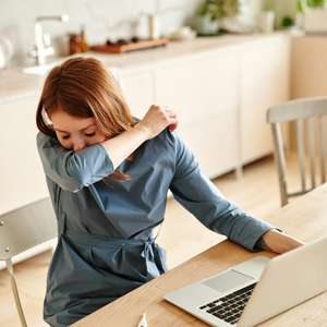 En kvinne sitter ved et bord med en PC, og snur seg vekk for å nyse i armkroken (Foto)