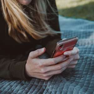 Kvinne ligger på et teppe på gresset i en park og ser på en mobil hun har i hendene (Foto)